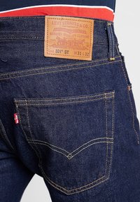 Detailní záběr na tmavě modré džíny Levi’s 501® ST, zadní kapsa s koženou nášivkou a červeným štítkem Levi's na švu kapsy.