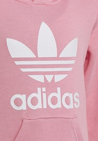 Felpa con cappuccio rosa chiaro, caratterizzata da un grande logo Adidas bianco e tre strisce bianche sul petto. Il materiale è liscio e texturizzato.