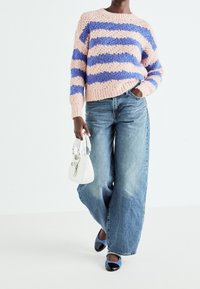 Personne portant un pull en maille épaisse rayé rose et bleu, un jean bleu à jambes larges, des chaussures bleues et tenant un petit sac blanc.