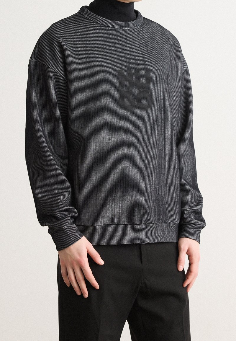 Sweat-shirt à rayures noires avec un col ras du cou, des poignets et un ourlet côtelés, présentant une texture de logo subtile sur le devant. Porté avec un pantalon foncé.