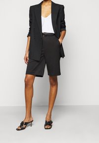 Blazer nero doppiopetto sopra un top bianco, abbinato a pantaloni shorts neri su misura e mule nere con tacco caratterizzati da un dettaglio a nodo.