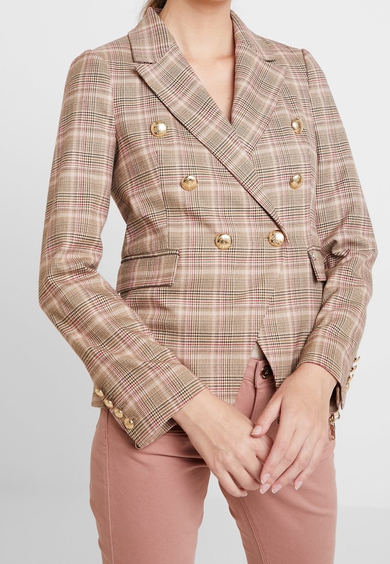 Femme portant un blazer beige à carreaux à double boutonnage avec des boutons dorés, par-dessus un pantalon rose pâle, les mains légèrement jointes devant.