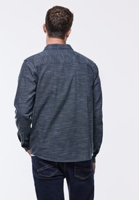 Camicia a maniche lunghe blu scuro con tessuto testurizzato, colletto a bottoni e orlo curvo, abbinata a jeans scuri, vista da dietro.