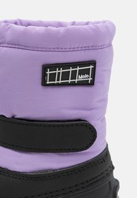 Botte en tissu violet avec semelle et sangle noires. Présente une surface texturée et une étiquette de marque avec un design en grille. Vue rapprochée de la partie supérieure.