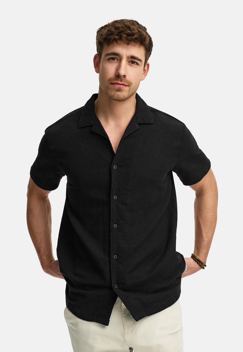Black short-sleeve shirt with a collared neckline, featuring subtle texture and buttons down the front. Worn over light-colored pants.