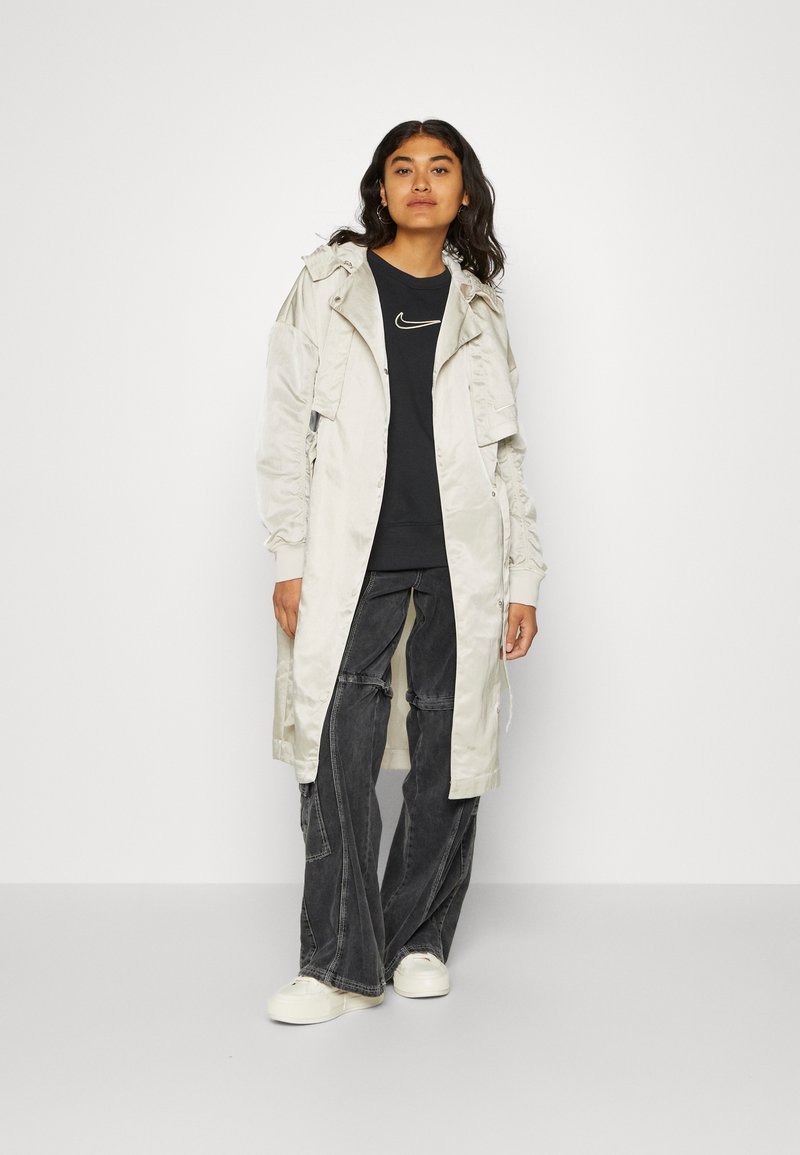 Mujer vestida con un abrigo largo beige claro, sudadera negra de Nike, pantalones oscuros de pierna ancha y zapatillas blancas, de pie contra un fondo sencillo.