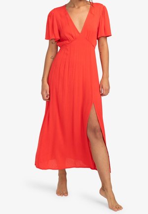 Femme portant une robe midi orange vif avec des manches volantées, un profond décolleté en V, une fente haute sur le côté, et pieds nus sur un fond blanc.