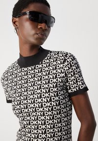 Zwart-wit gebreide t-shirt met een herhalend "DKNY" logo patroon, geribbelde zwarte kraag, korte mouwen en een aansluitende pasvorm.