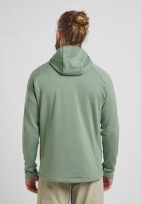 Hoodie long à manches longues, en vert texturé, avec une capuche attachée, présentant un design ajusté et des manches raglan, adapté pour le sport.