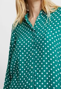 Chemise boutonnée bleu sarcelle avec des pois blancs, un col, une coupe décontractée et une texture lisse. Les boutons sont assortis en bleu sarcelle.