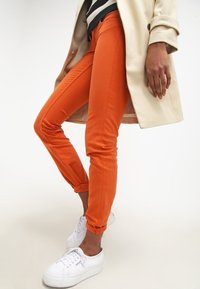 Orange slim-fit byxor med uppkavlade vristerna, tillverkade av ett glatt tyg, kombinerade med vita sneakers och en ljusfärgad kappa.