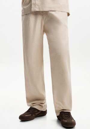 Beige loose-fitting pants worn with dark brown casual sneakers on a white background.