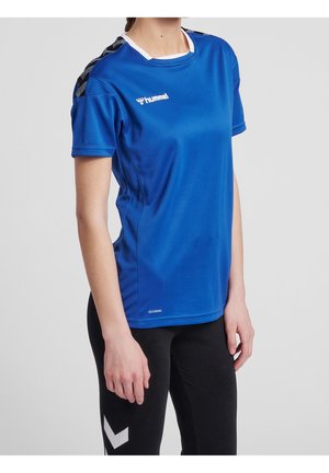 Person, die ein blaues, kurzärmliges Hummel-Sportshirt und schwarze Hose mit weißem Chevron-Muster auf hellem, einfarbigem Hintergrund trägt.