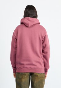 Mikina s kapucňou v tlmené mauve farbe, s klokankou a sťahovacou šnúrkou na kapucni, skombinovaná s voľnými zeleno-hnedými nohavicami.