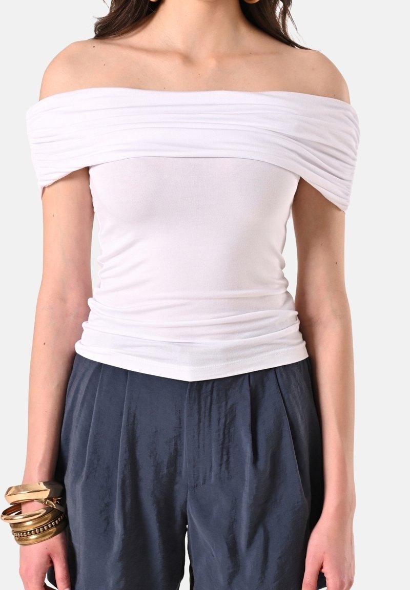 Haut blanc à épaules dénudées avec une texture froncée, un corsage ajusté et une finition lisse. Porté avec un pantalon ample, bleu foncé, texturé.