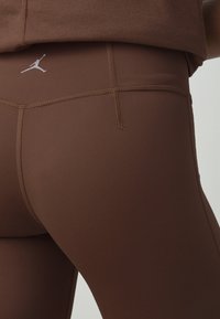 Gros plan sur des leggings de sport marron avec des coutures visibles et un petit logo Jumpman blanc sur la partie supérieure de la hanche.