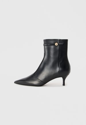 Black leather ankle boot with a pointed toe and a small, tapered heel. Features a decorative strap with a gold button accent. Smooth texture.