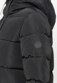 Schwarze Puffjacke mit gestepptem Design, ausgestattet mit einer Kapuze, strukturiertem Material und einem runden LOGO-Patch auf dem linken Ärmel.