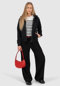 Felpa nera con zip, camicia a righe bianche e nere, pantaloni larghi neri, cintura rossa, borsa rossa e calzature nere su uno sfondo grigio.