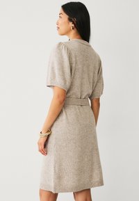 Gehaakte jurk in lichtbeige met korte pofmouwen, een tailleband en een lengte tot de knie. Zachte textuur en casual ontwerp, minimale details.