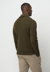 Olivgrüner, gerippter Strickpullover mit umgeschlagenem Kragen, kombiniert mit hellbraunen, strukturierten Hosen. Die Rückansicht zeigt ein tailliertes Design.