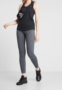 Kobieta ubrana w czarny bezrękawnik Reebok, szare legginsy, czarne sneakersy oraz czarną torbę na ramię Reebok, idąca na białym tle.