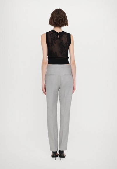 Débardeur en dentelle noire avec un dos en forme de trou de serrure, associé à un pantalon gris clair ajusté avec poches arrière, jambe droite et pli marqué.