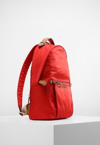 Rød nylon rygsæk med en glat overflade, buet design, brune læderdetaljer og en frontlomme med lynlås. Har polstrede skulderstropper.