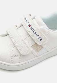 Sneaker sintetica bianca con cinturini in Velcro, caratterizzata da una superficie testurizzata, interno blu chiaro e un logo sul cinturino.