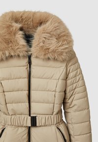 Doudoune beige avec un col en fausse fourrure marron duveteuse, fermeture éclair, taille ceinturée, texture matelassée et poches latérales.