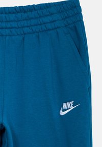 Tirkīzzilas sporta bikses ar elastīgu jostu, kurām kreisajā pusē ir balts izšūts Nike logo un mīksta, tekstūrēta auduma. 