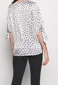 Witte satijnen blouse met zwarte en beige stippen, met ellebooglange mouwen die in knopen zijn vastgebonden, gedragen met zwarte broeken, gezien van de achterkant.