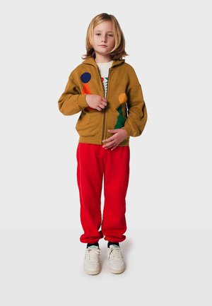 Brauner Zip-Hoodie mit bunten Kreis- und Dreieck-Patches, kombiniert mit roten Jogginghosen und weißen Turnschuhen mit grünen Akzenten.