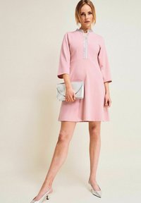 Rosa klänning med ett åtsittande liv och utsvängd kjol, med en hög halsringning prydd med silverdekorationer. Matchad med en silverklutch.