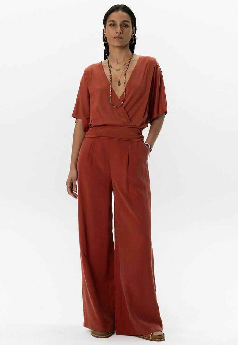 Combinaison en terracotta avec un décolleté en V profond et des manches courtes. Présente des pantalons évasés et une taille ceinturée pour une silhouette structurée.