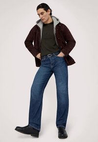 Brun mockajacka över en grå hoodie, grön t-shirt med logotyp, blå jeans med raka ben och svarta ankelstövlar. Enkel design och avslappnad passform.