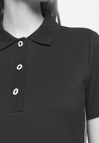 Närbild av en person som bär en svart strukturerad polo med tre vita knappar och korta ärmar.