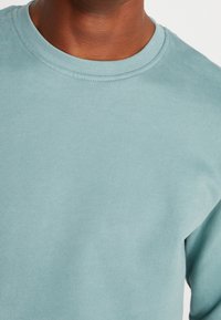 Sudadera azul claro con cuello redondo y ribete acanalado. Textura lisa y ajuste ligeramente holgado. Sin patrones ni gráficos visibles.