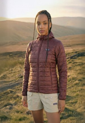 Vrouw met gevlochten haar staat buiten in een bruine doorgestikte jas en beige korte broek met heuvels op de achtergrond tijdens zonsondergang.