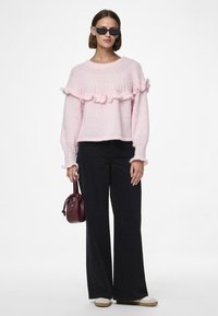 Pull en peluche rose avec des détails volants, manches longues et col côtelé. Associé à un pantalon noir à jambe large et un petit sac à main bordeaux.