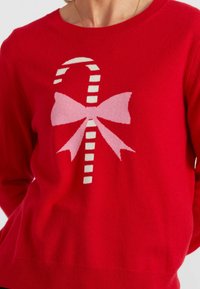 Pull en tricot rouge avec un design de sucre d'orge et un nœud rose. Texture douce avec un col rond et un ourlet côtelé.