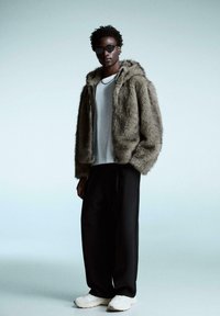 Jachetă din blană artificială de culoare maronie cu glugă, purtată peste o cămașă gri, asortată cu pantaloni largi negri și adidași albi. Modelul poartă ochelari de soare negri.