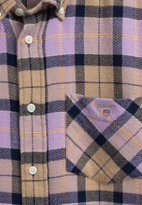 Koszula z flaneli w kratę w kolorach beżowym, lawendowym i granatowym. Posiada zapięcie na guziki, kołnierzyk oraz naszywaną kieszeń z zaakcentowanym wzorem i logo.