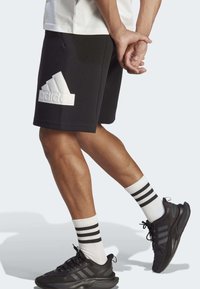 Musta urheilushortsit, joissa on valkoinen koholla oleva Adidas-logo, yhdistettynä mustiin kenkiin ja valkoisiin sukkiin, joissa on mustat raidat. Sujuva kangastuntuma.