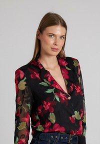Blusa floral con un tejido negro transparente adornado con patrones de flores rojas y verdes, un cuello con muescas y un único botón decorativo.