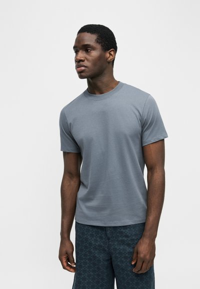 Άνδρας που στέκεται με το σώμα στραμμένο αριστερά, φορώντας απλό γκρι κοντομάνικο μπλουζάκι και σκούρα σορτς με σχέδια, μπροστά σε απλό λευκό φόντο.