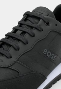 Czarny sneaker z teksturowaną tkaniną i gładką nakładką, wyposażony w płaskie sznurowadła, gumową podeszwę oraz delikatne oznaczenia po boku.