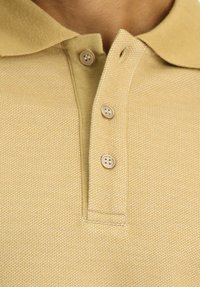 Beige Poloshirt aus strukturiertem Stoff, mit klassischem Kragen und drei beigen Knöpfen am Hals.