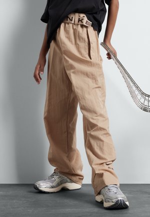 Person iført beige bukser med vide ben og et synligt Nike-bælte, sort skjorte og grå sneakers, der holder i håndtaget på en metallic trådkurv.