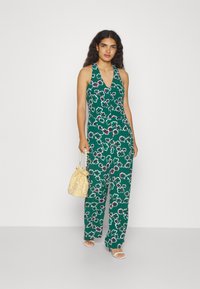 Grønn floral jumpsuit med halterneck-design, prydet med hvite og rosa blomster, løs passform, kombinert med en lys gul, teksturert veske.